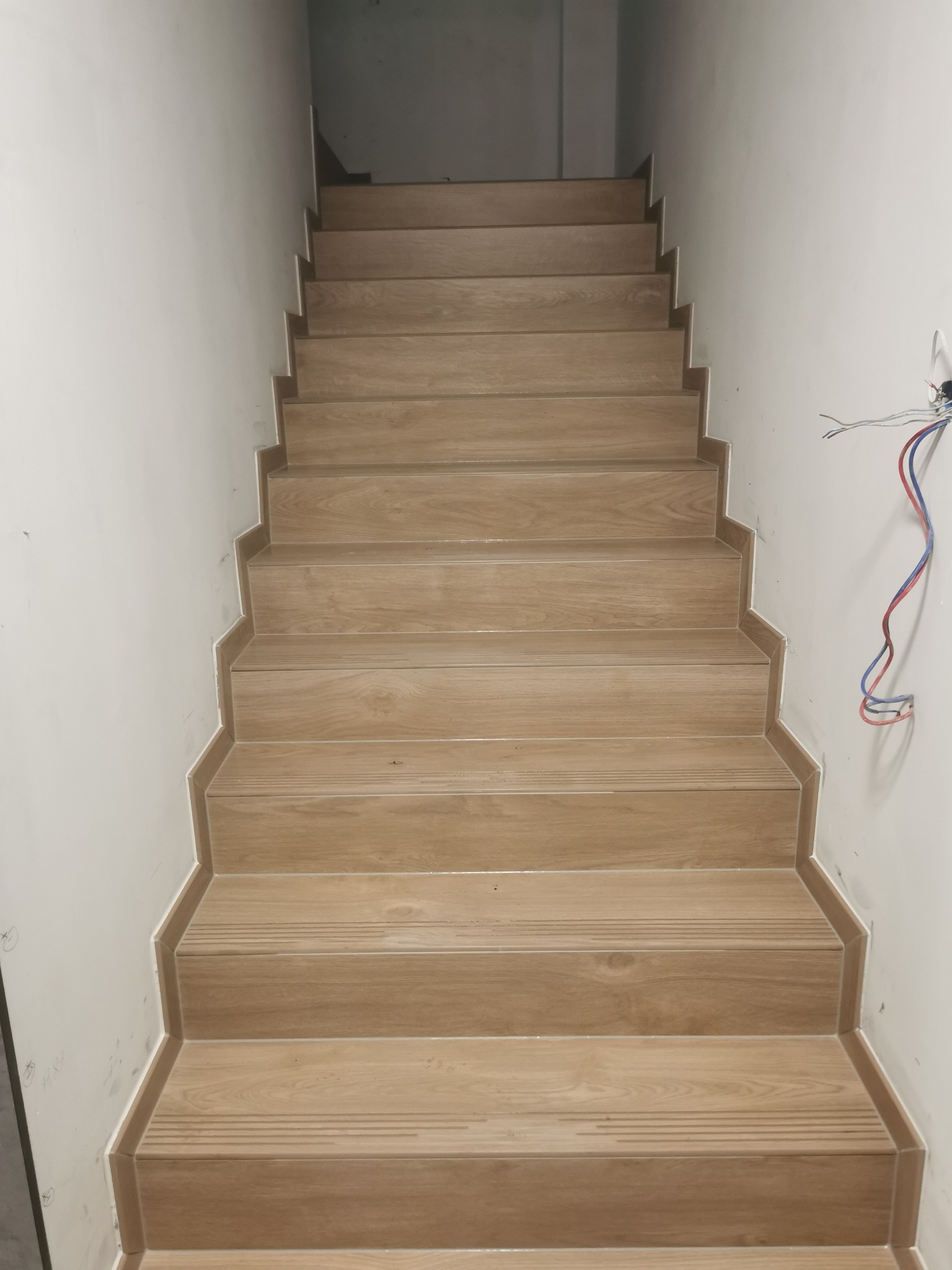 Wykończone schody z płytek imitujących drewno, prowadzące w górę. Widoczne detale wykończenia krawędzi i fragment instalacji elektrycznej na ścianie.