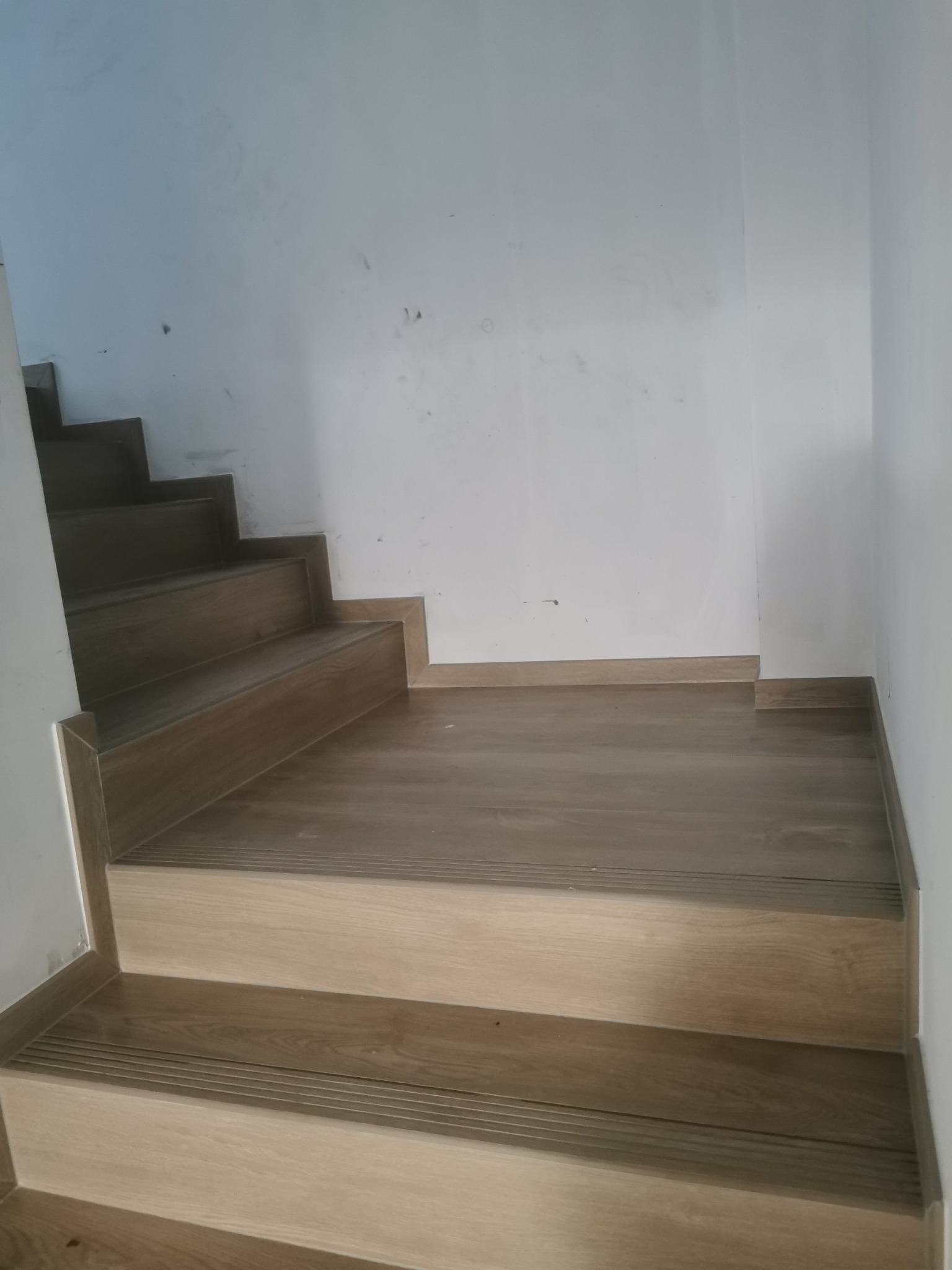 Wykończone schody wewnętrzne z drewnopodobnych paneli, z listwami przypodłogowymi. Widok z dołu, ściany białe, surowe.