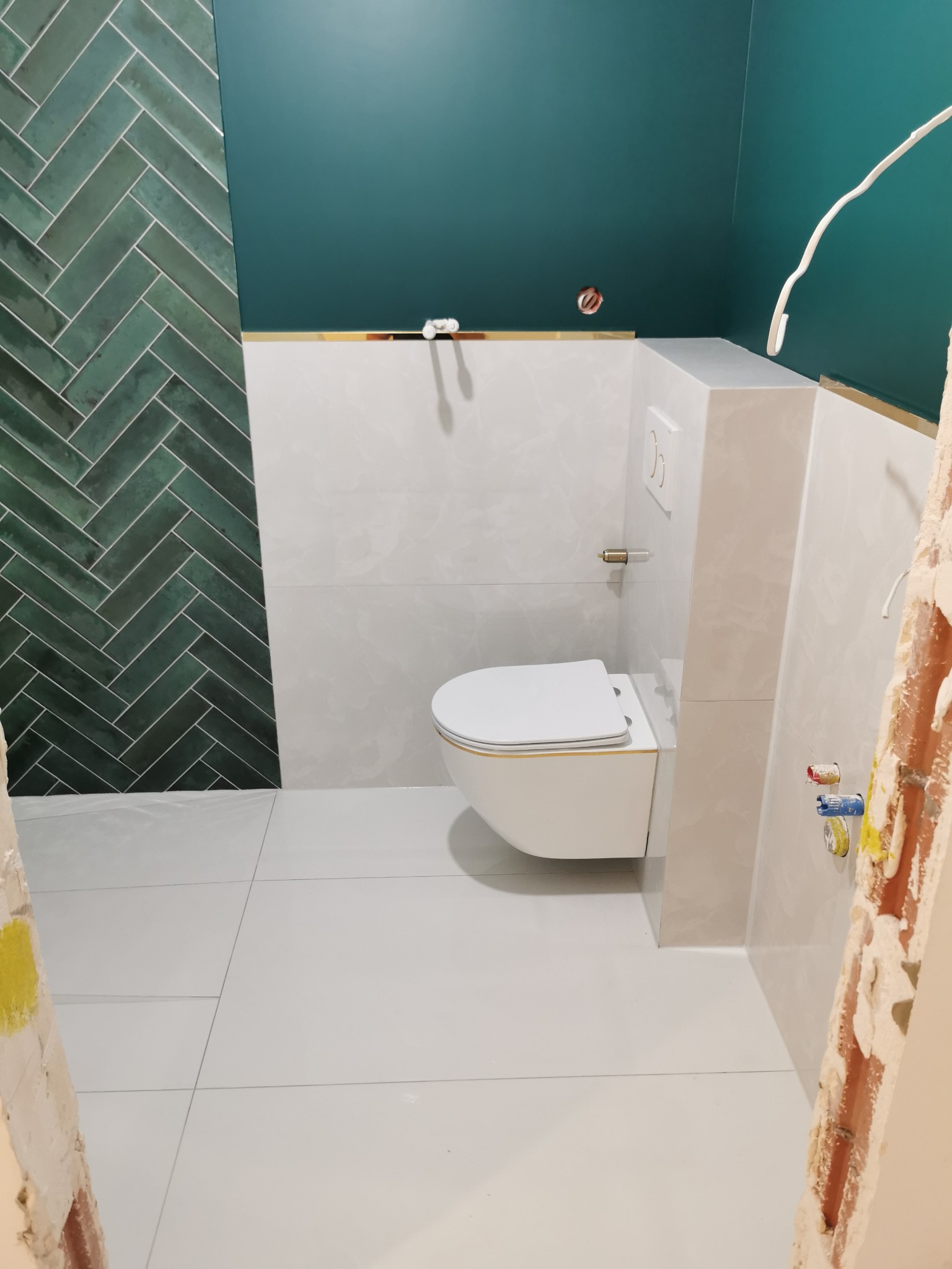 Nowoczesna łazienka w trakcie wykończenia: białe płytki, zielona ściana z wzorem jodełki, podwieszana toaleta. Widoczne elementy instalacji i niedokończone wykończenia.