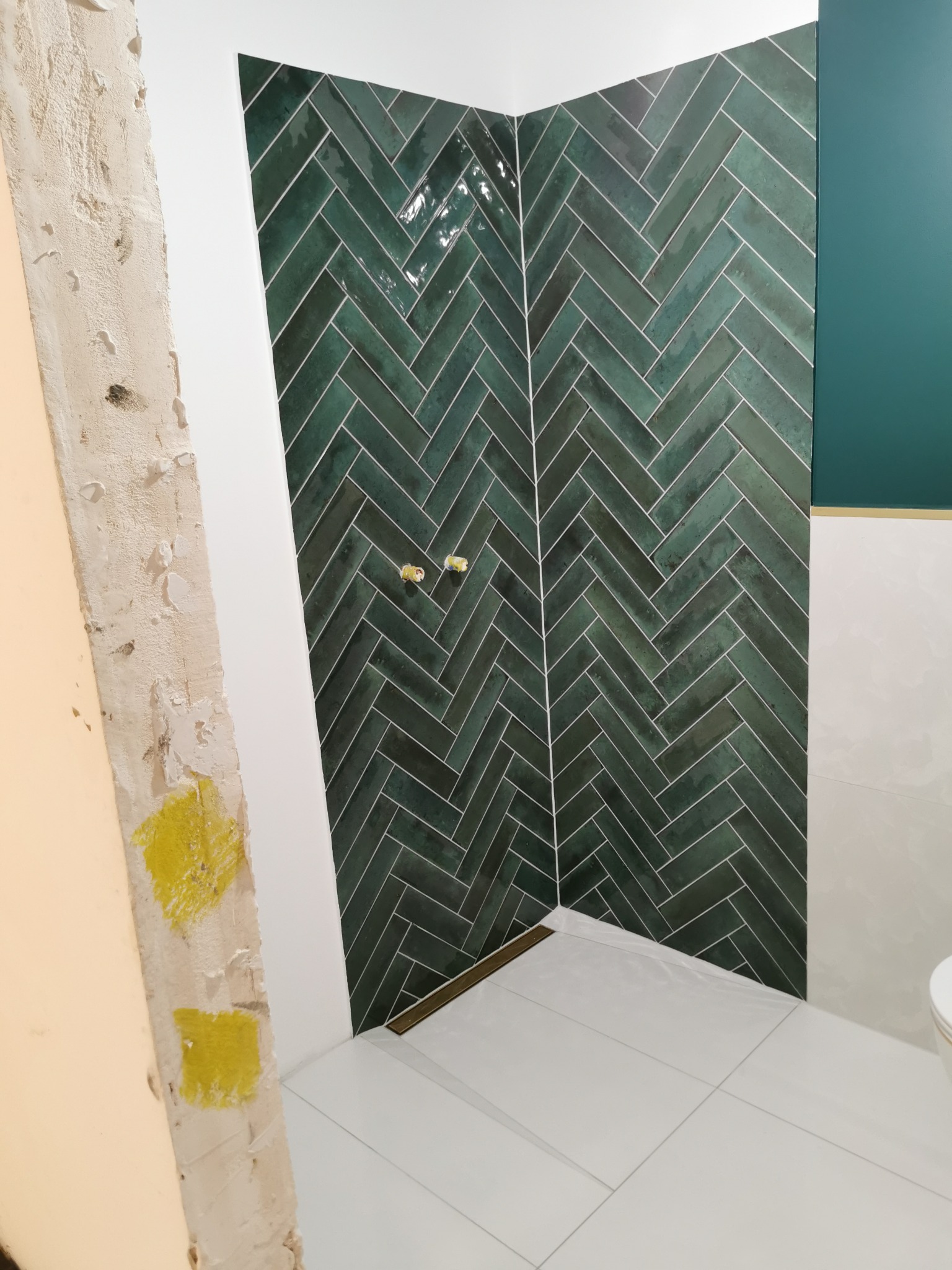 Wykończona ściana kabiny prysznicowej z zielonych płytek ułożonych w jodełkę, biała podłoga i fragment ściany w trakcie prac wykończeniowych.