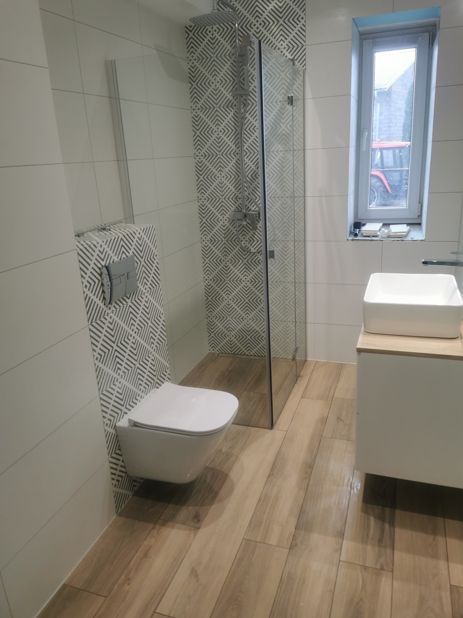 Nowoczesna łazienka po remoncie: białe płytki, drewnopodobna podłoga, kabina prysznicowa ze wzorem, podwieszana toaleta i umywalka nablatowa. Widok na okno.