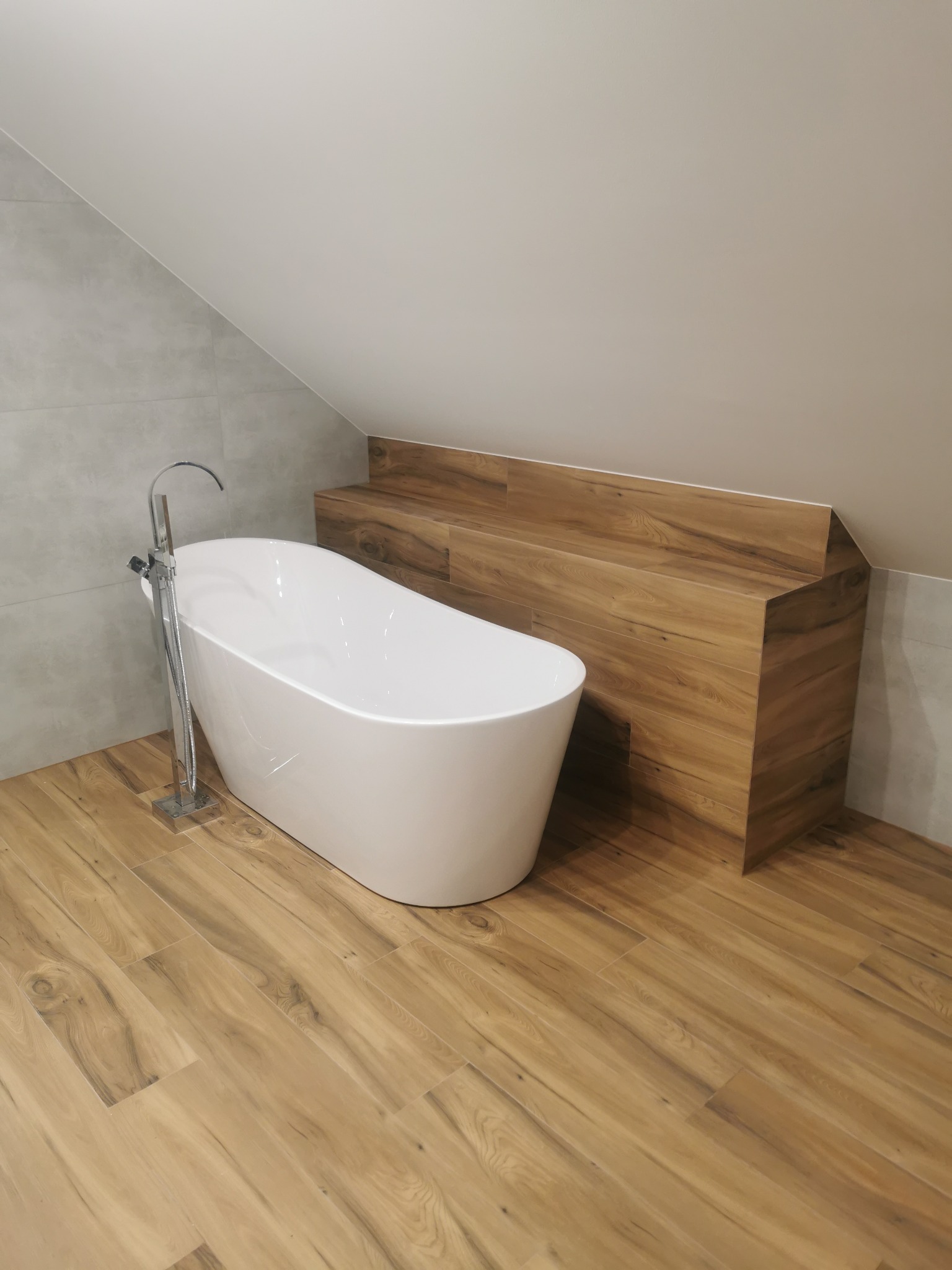 Nowoczesna łazienka na poddaszu z wolnostojącą wanną, drewnopodobną zabudową i płytkami imitującymi drewno na podłodze. Minimalistyczny design.