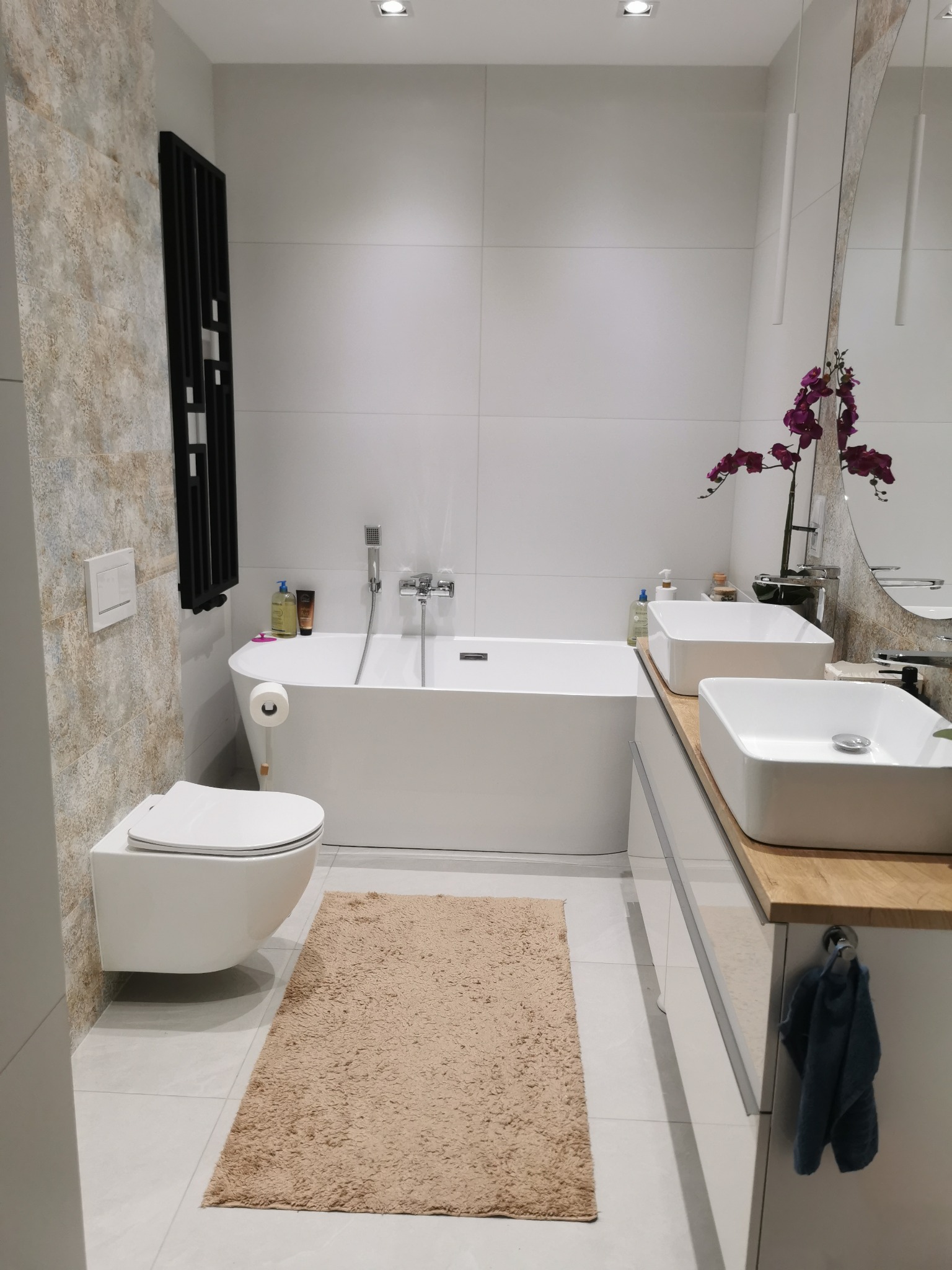 Nowoczesna łazienka z wolnostojącą wanną, podwieszaną toaletą i dwiema umywalkami nablatowymi. Uwagę zwraca dekoracyjna ściana z efektem kamienia i minimalistyczne oświetlenie.