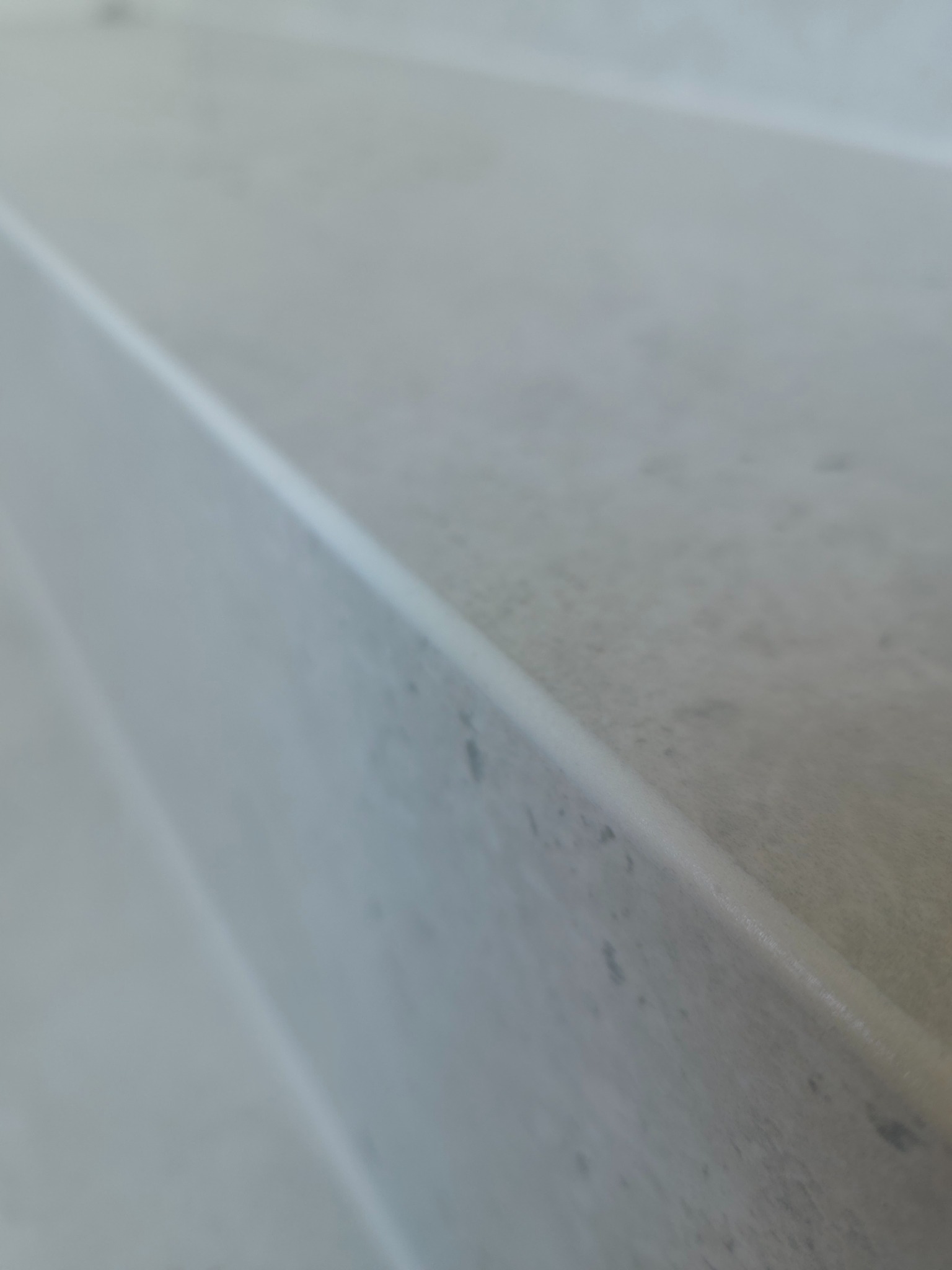 Szczegółowe ujęcie krawędzi jasnoszarej płytki, prawdopodobnie ceramicznej, z delikatną fakturą. Widoczne subtelne przebarwienia i zaokrąglona krawędź.