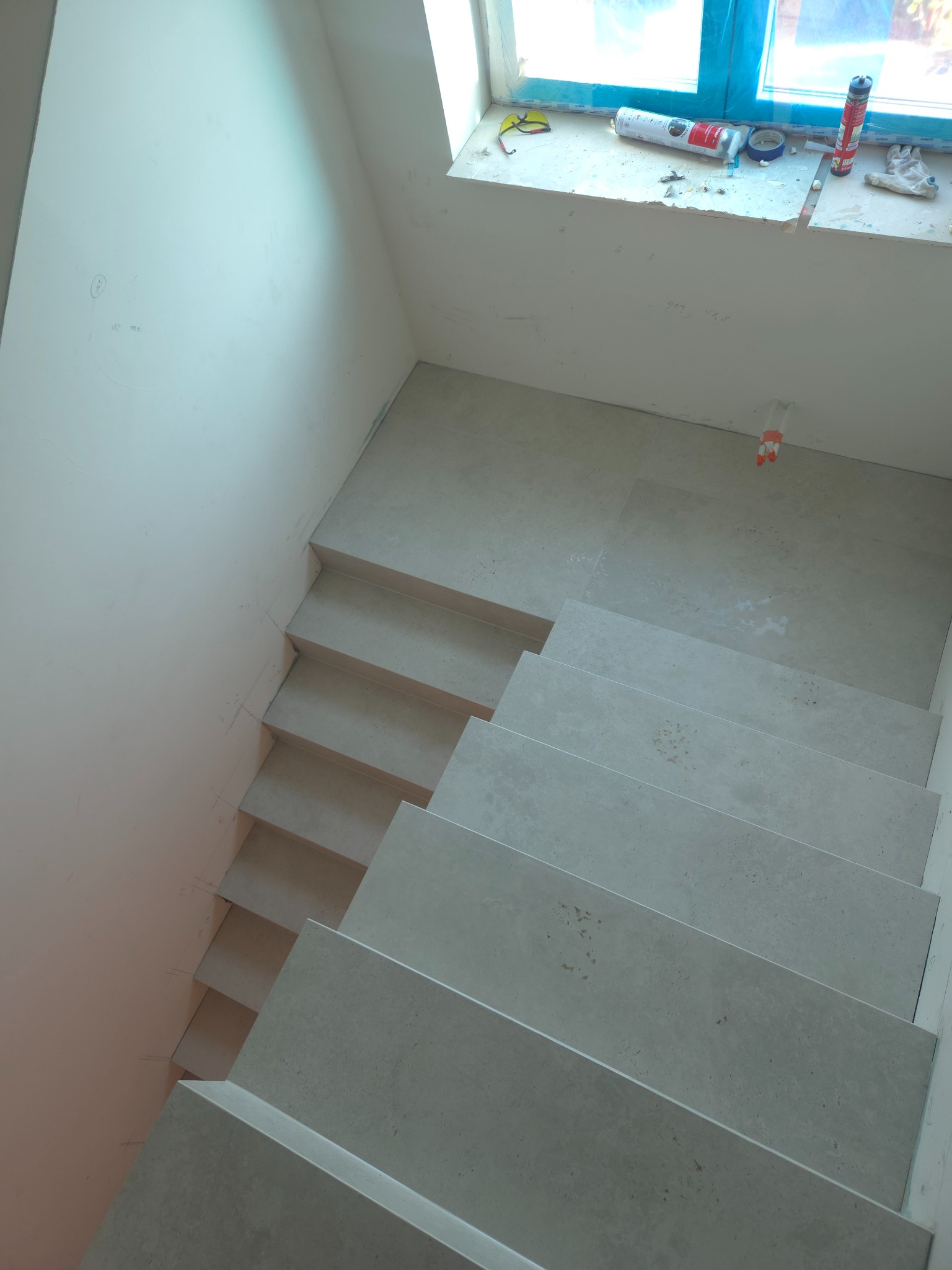 Wykończone schody wewnętrzne z szarych płytek, widok z góry. Na parapecie okna widoczne narzędzia i materiały budowlane. Minimalistyczny design.