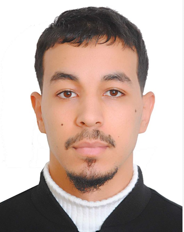 Portret młodego mężczyzny z brązowymi oczami i ciemnymi włosami, ubrany w biały golf i czarną marynarkę, na białym tle.