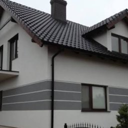 Grzegorz Łazarski - Dwukondygnacyjny dom z szarą, poziomą dekoracją elewacji i ciemną dachówką. Widoczne okna z brązowymi ramami i mały balkon. Ujęcie z poziomu gruntu.