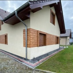 Grzegorz Łazarski - Dom jednorodzinny z beżową elewacją i drewnopodobnymi panelami. Widoczne okna z roletami, rynna i fragment posesji z kostką brukową i trawnikiem.