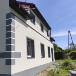 Grzegorz Łazarski - Odnowiona elewacja domu z szarymi detalami: narożnik z pasami, obramowania okien i cokół. Widok z boku, błękitne niebo w tle.