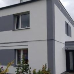 Grzegorz Łazarski - Dwukondygnacyjny dom z jasną elewacją i szarymi akcentami. Widoczne okna i wejście. Minimalistyczny design z roślinnością w tle.