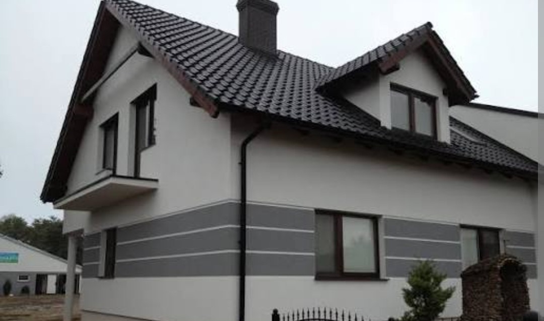 Dwukondygnacyjny dom z szarą, poziomą dekoracją elewacji i ciemną dachówką. Widoczne okna z brązowymi ramami i mały balkon. Ujęcie z poziomu gruntu.