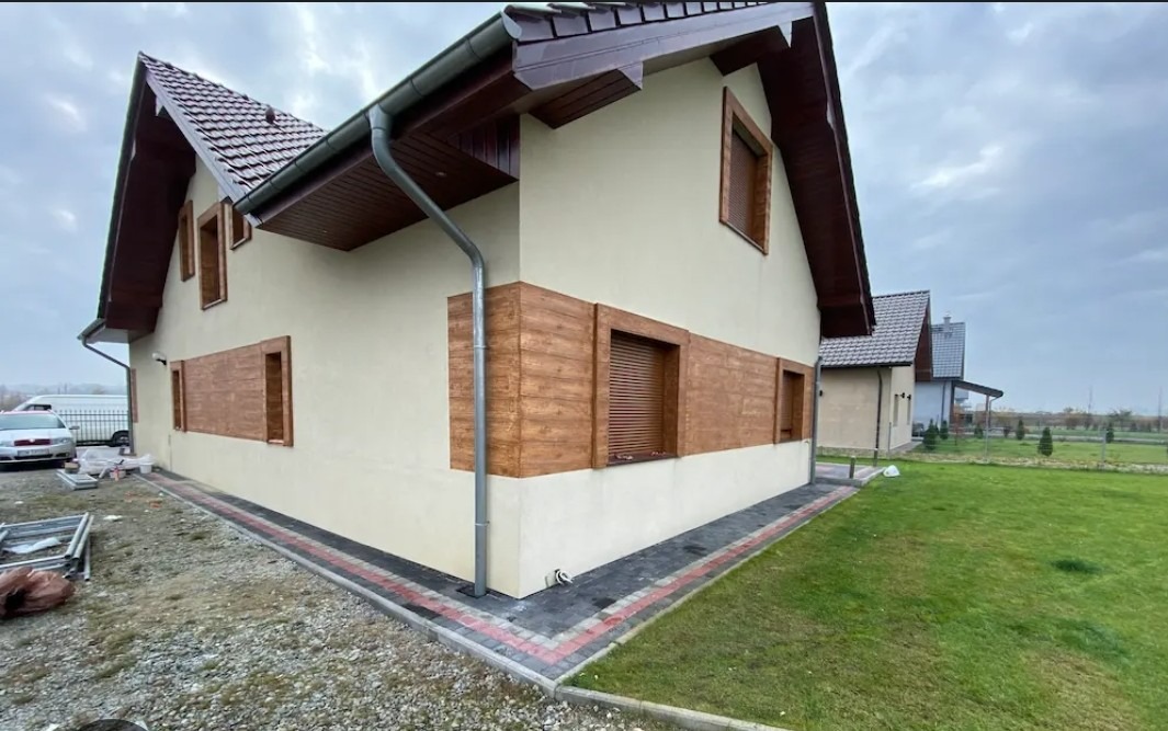 Dom jednorodzinny z beżową elewacją i drewnopodobnymi panelami. Widoczne okna z roletami, rynna i fragment posesji z kostką brukową i trawnikiem.