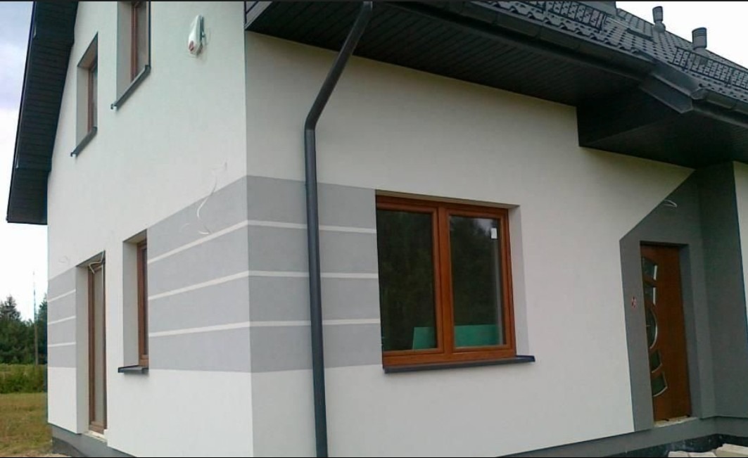 Dwukondygnacyjny dom z jasną elewacją i szarymi pasami, brązowe okna, ciemny dach i rynna. Widok z boku, zieleń w tle.