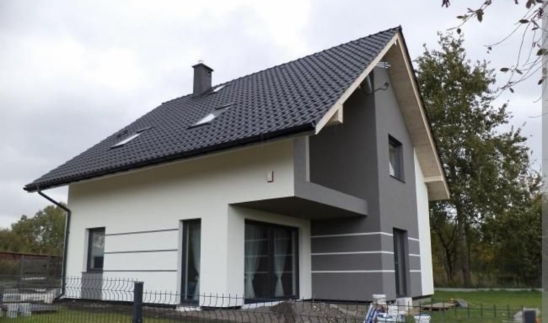 Dwukolorowa elewacja domu jednorodzinnego z ciemną dachówką i szarymi akcentami. Widoczne okna dachowe i fragment ogrodzenia. Nowoczesny design.