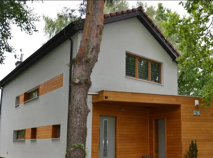 Nowoczesny dom z szarą elewacją i drewnianymi akcentami, wkomponowany w zieleń. Widoczny fragment pnia drzewa przy elewacji i drewniane panele pod oknami.