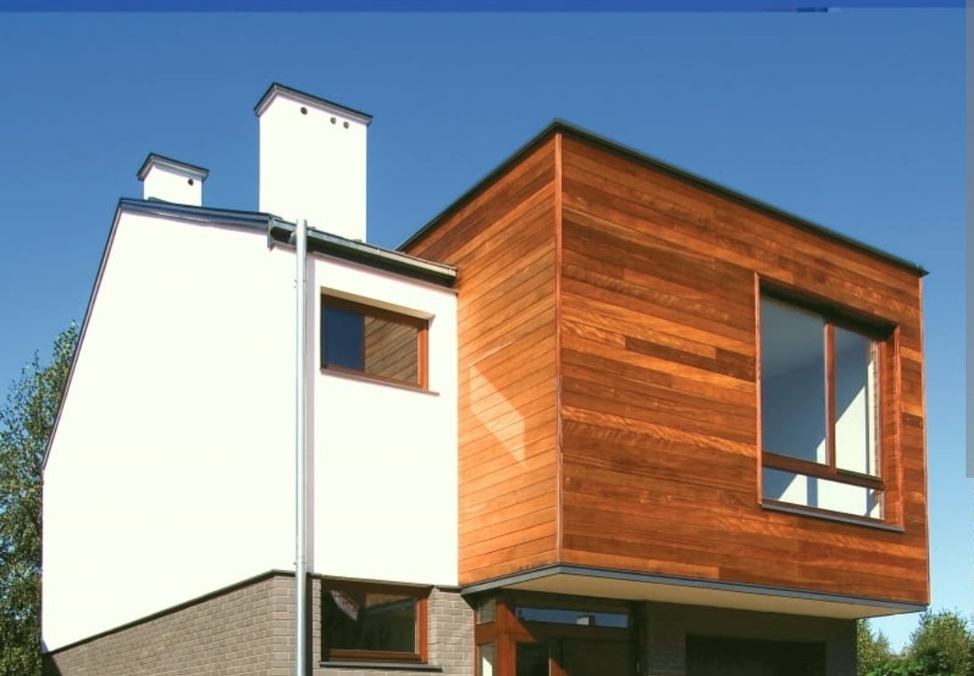 Nowoczesny dom z elewacją łączącą biały tynk i drewniane panele. Widoczne okna z drewnianą stolarką i fragment zieleni. Jasne, słoneczne oświetlenie.