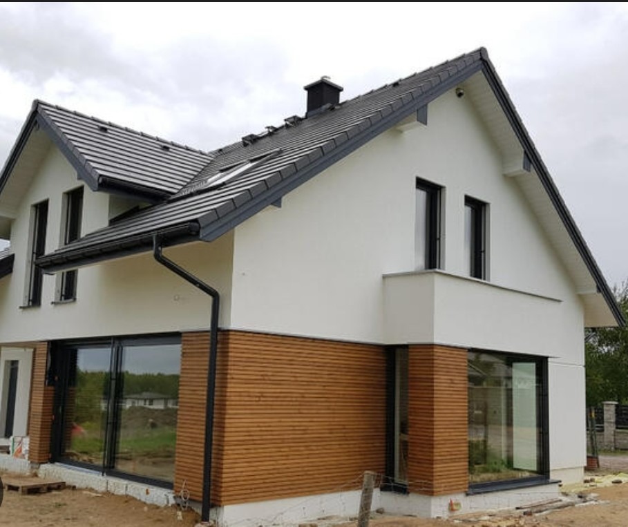 Nowoczesny dom jednorodzinny z białą elewacją i drewnianą okładziną. Duże okna z czarnymi ramami. Dach pokryty ciemną dachówką. Widok z zewnątrz, w tle zieleń.