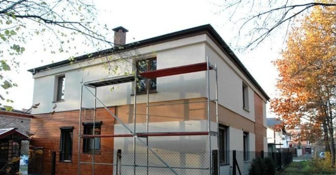 Dwukondygnacyjny dom w trakcie prac. Część ściany drewniana, część biała. Widoczne rusztowania i fragmenty drzew. Ujęcie z dołu, w tle zabudowa jednorodzinna.