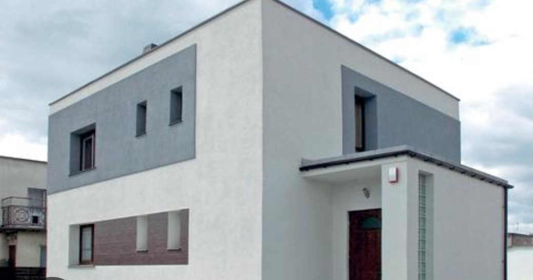 Nowoczesny dom z białą elewacją i szarymi akcentami wokół okien, widoczny z perspektywy narożnej, z ciemnymi ramami okiennymi i drzwiami wejściowymi.