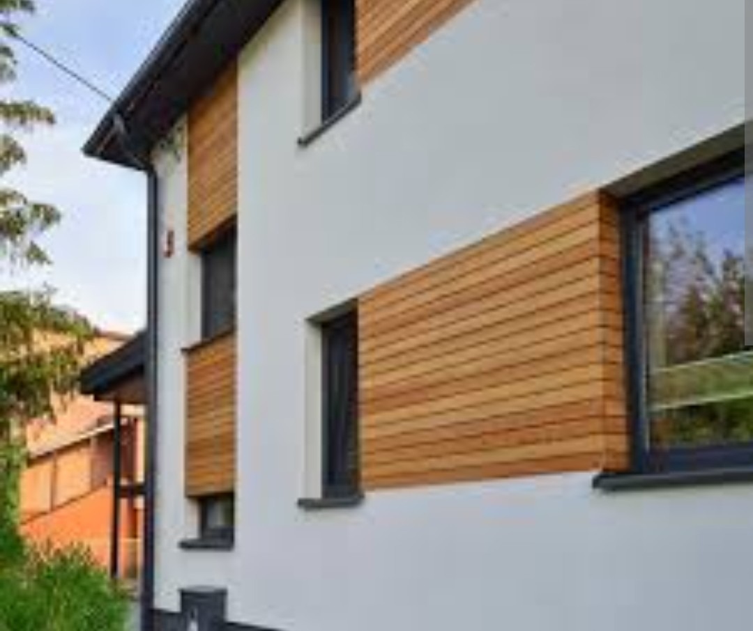 Nowoczesna elewacja domu w Toruniu z białym tynkiem i drewnianymi panelami. Widoczne okna z czarnymi ramami i fragment dachu. Elegancki design.