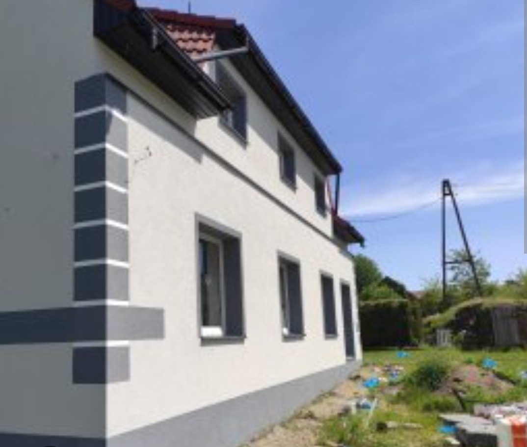 Odnowiona elewacja domu z szarymi detalami: narożnik z pasami, obramowania okien i cokół. Widok z boku, błękitne niebo w tle.