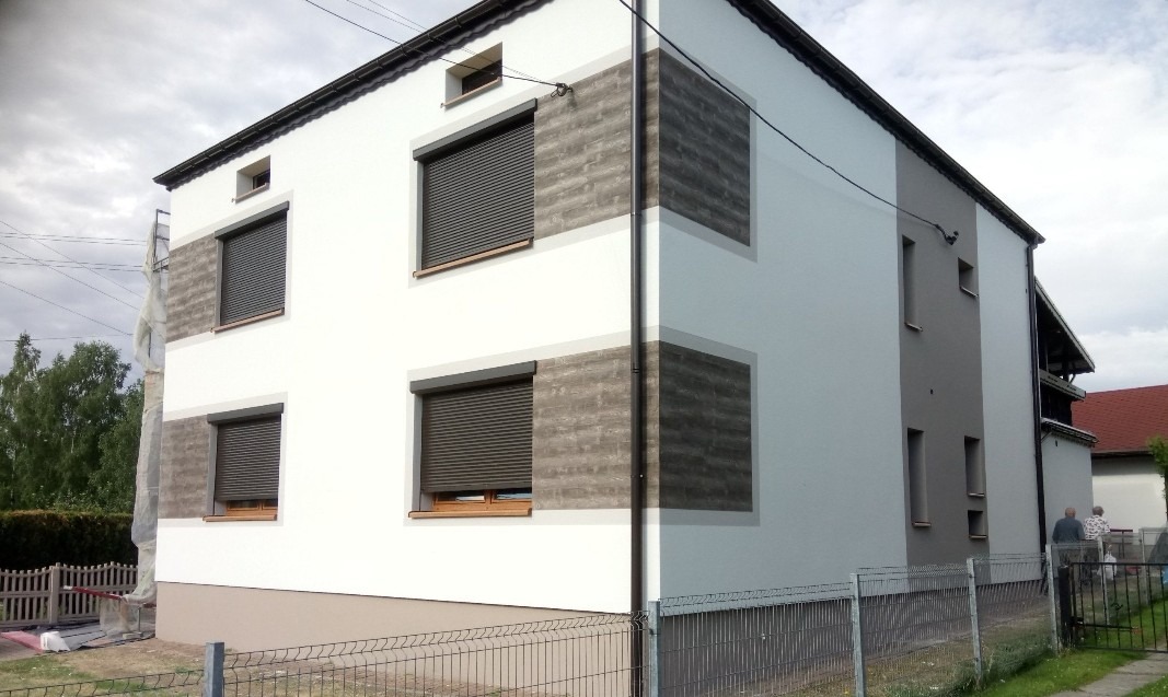 Dwukondygnacyjny dom z biało-szarą elewacją, z roletami w oknach. Fragment elewacji z fakturą betonu. Widoczne ogrodzenie i zieleń w tle. Dwie osoby w oddali.