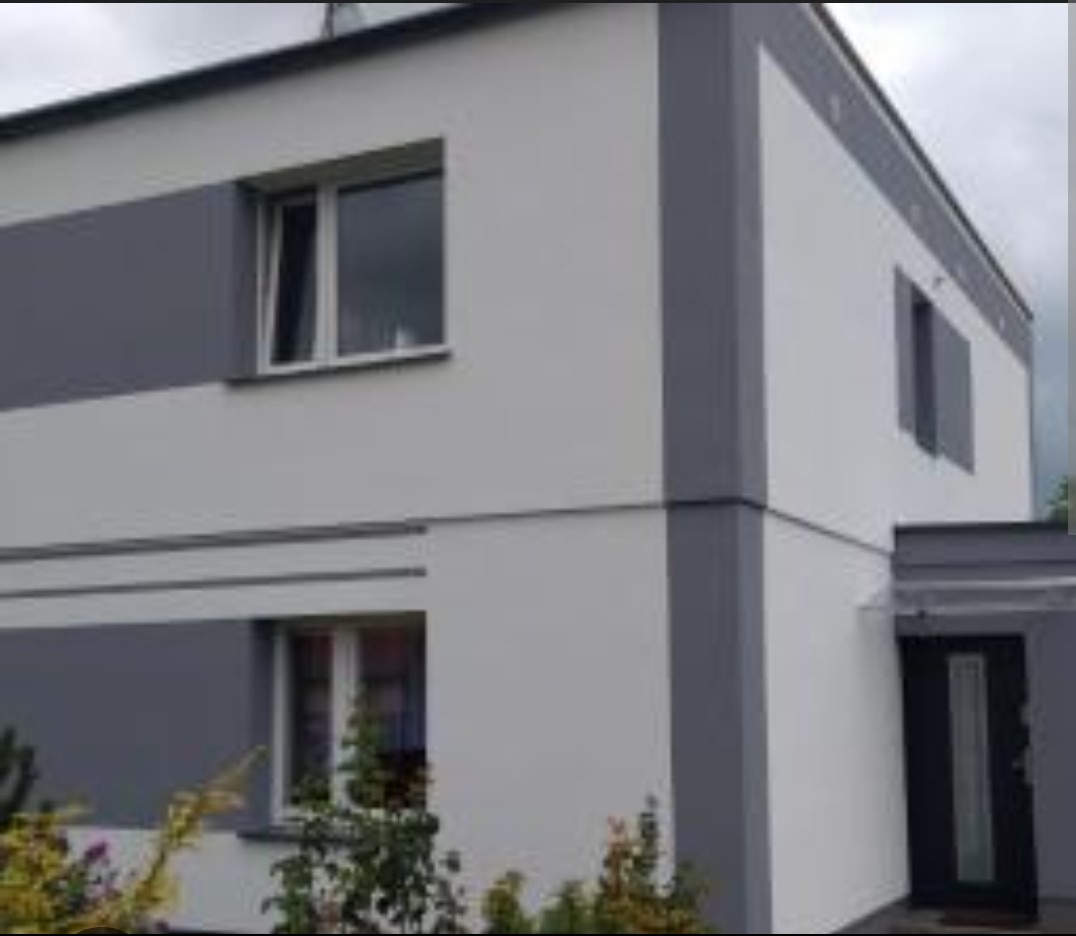 Dwukondygnacyjny dom z jasną elewacją i szarymi akcentami. Widoczne okna i wejście. Minimalistyczny design z roślinnością w tle.