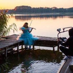 REELS STUDIO PATRYK BIAŁAS - Kamerzysta filmuje młodą kobietę w błękitnej sukni grającą na skrzypcach na drewnianym pomoście nad jeziorem o zachodzie słońca. Ujęcie z łodzi, widoczny sprzęt filmowy.