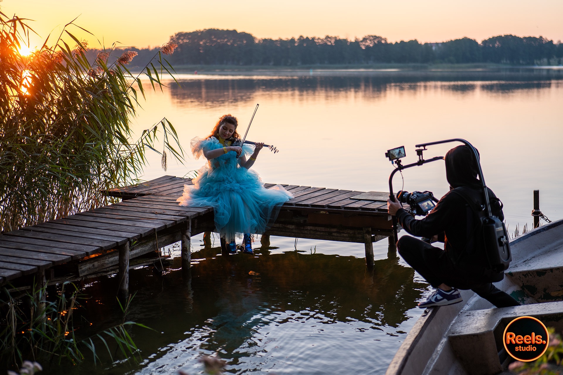 Kamerzysta filmuje młodą kobietę w błękitnej sukni grającą na skrzypcach na drewnianym pomoście nad jeziorem o zachodzie słońca. Ujęcie z łodzi, widoczny sprzęt filmowy.
