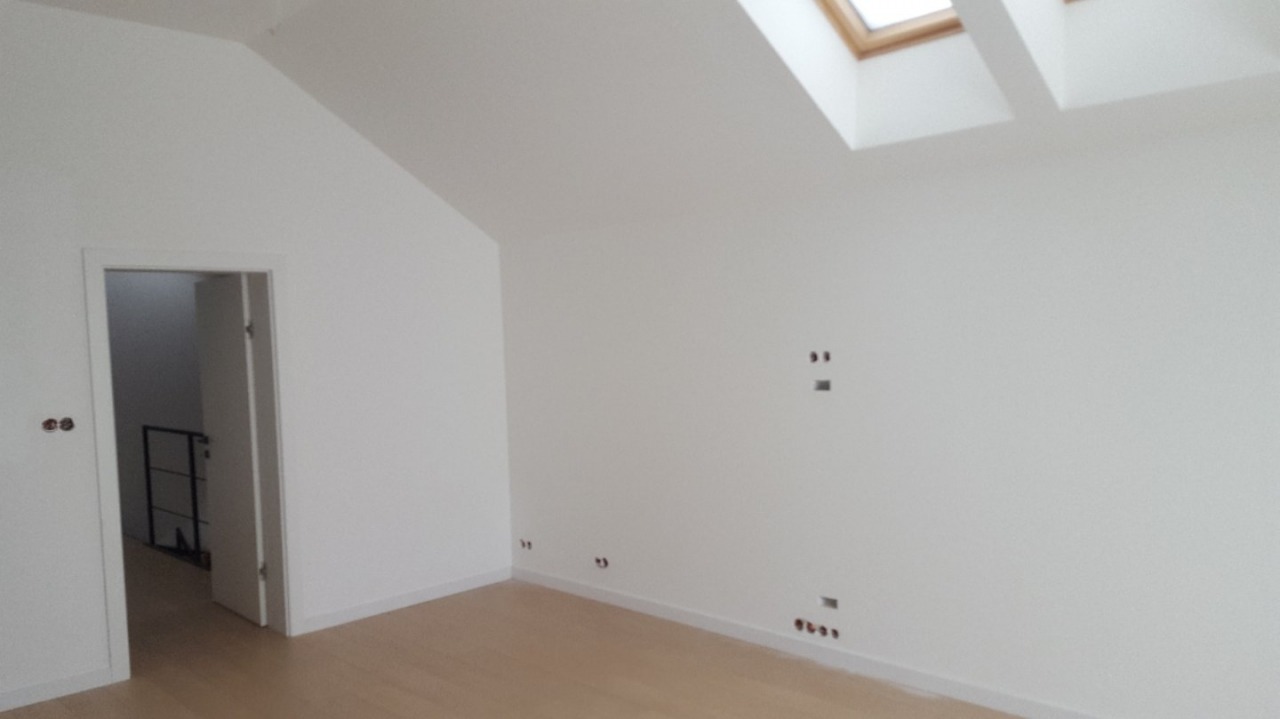 Jasne wnętrze poddasza po remoncie: białe ściany, drewniana podłoga, okno dachowe i otwarte drzwi do kolejnego pomieszczenia z czarną balustradą.