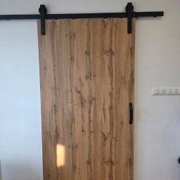 ADMIN ANNA LAUDAŃSKA - Drewniane drzwi przesuwne w stylu rustykalnym z czarnym, metalowym systemem zawieszenia. Widoczne detale faktury drewna i minimalistyczny uchwyt.