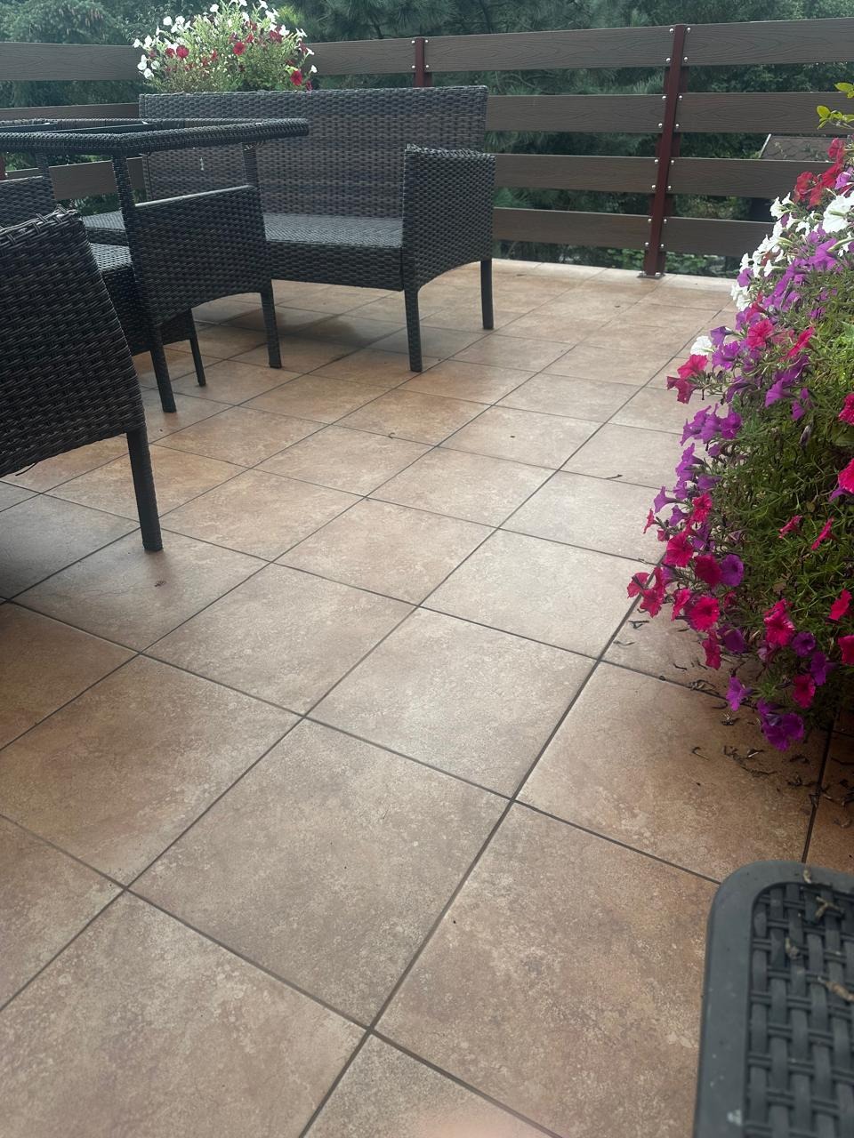 Odnowiony taras z beżowymi płytkami, rattanowe meble ogrodowe i kwiaty w donicach. Widok na drewniane balustrady i zieleń w tle. Jasne, naturalne światło.