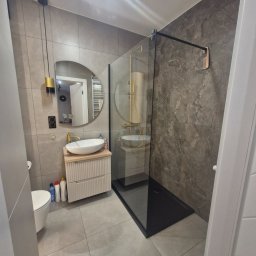 Monika Wierczyńska - Nowoczesna łazienka po remoncie: szare płytki, kabina prysznicowa typu walk-in z czarnym brodzikiem, okrągłe lustro nad umywalką na szafce z frezowanymi frontami.