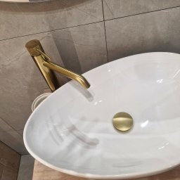 Monika Wierczyńska - Biała, owalna umywalka nablatowa z złotą armaturą i korkiem, na tle szarych płytek imitujących marmur. Nowoczesna łazienka po remoncie.