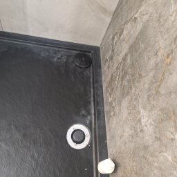 Monika Wierczyńska - Czarny brodzik z odpływem i kosmetykami przy marmurowej ścianie. Widoczne ślady użytkowania, wymagające czyszczenia. Ujęcie z góry, bliski kadr.