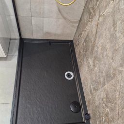 Monika Wierczyńska - Nowoczesny, czarny brodzik kabiny prysznicowej z odpływem, otoczony marmurowymi płytkami. Złoty uchwyt prysznica w tle. Minimalistyczny design łazienki.