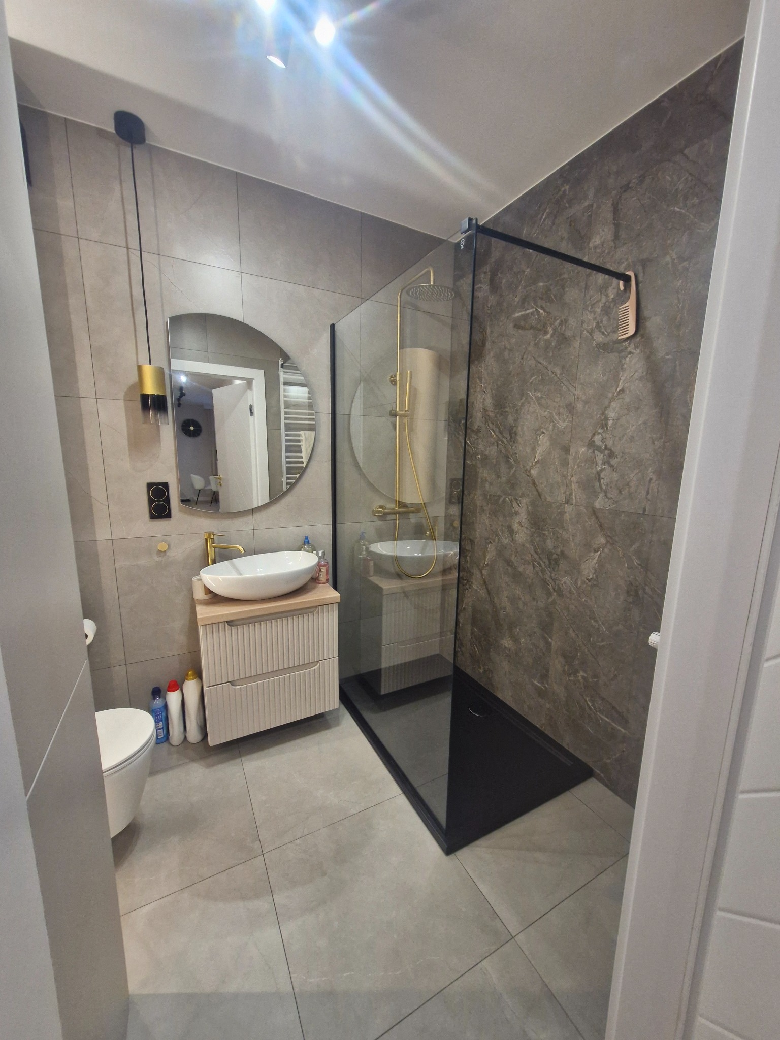 Nowoczesna łazienka po remoncie: szare płytki, kabina prysznicowa typu walk-in z czarnym brodzikiem, okrągłe lustro nad umywalką na szafce z frezowanymi frontami.