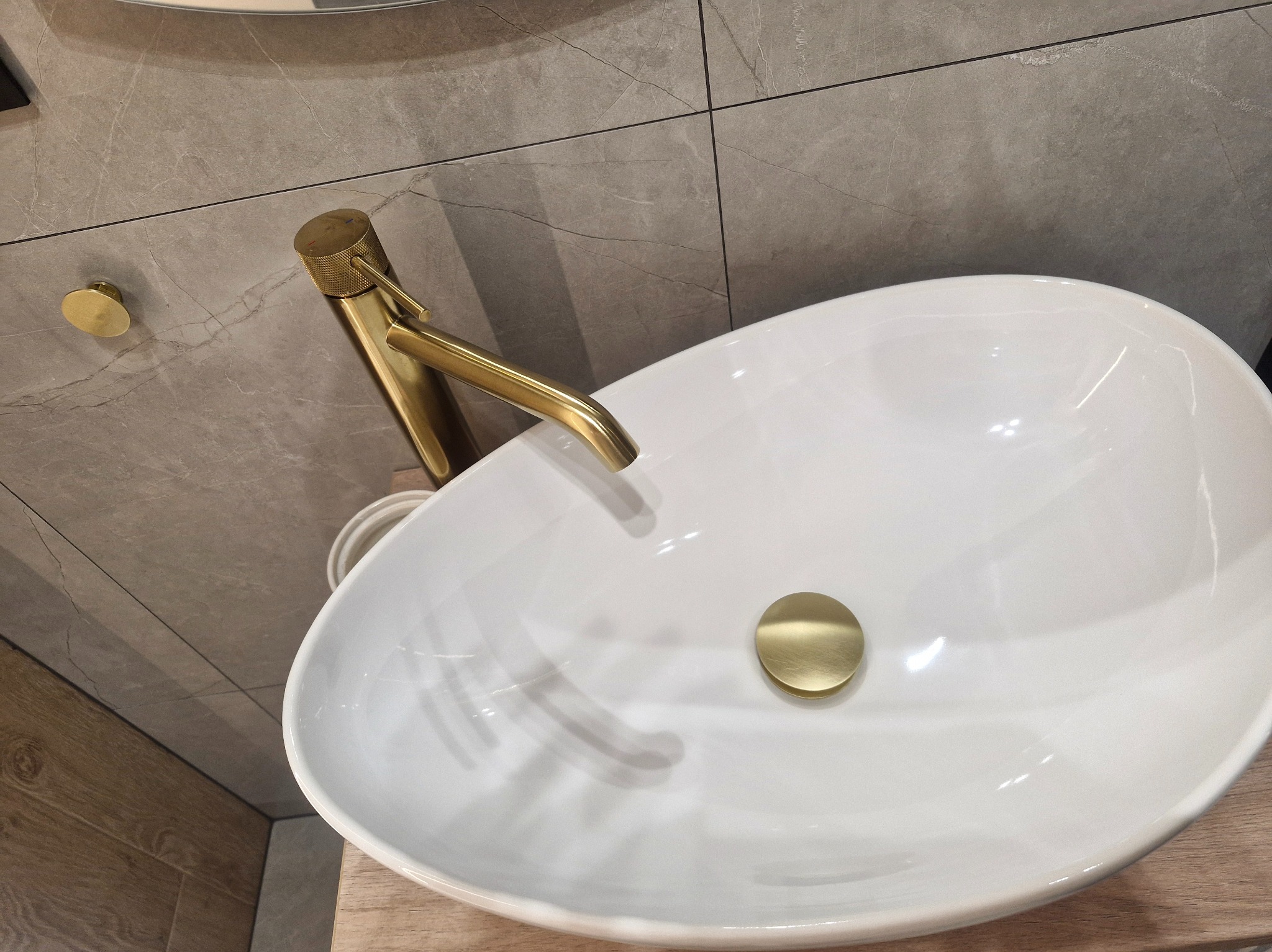 Biała, owalna umywalka nablatowa z złotą armaturą i korkiem, na tle szarych płytek imitujących marmur. Nowoczesna łazienka po remoncie.