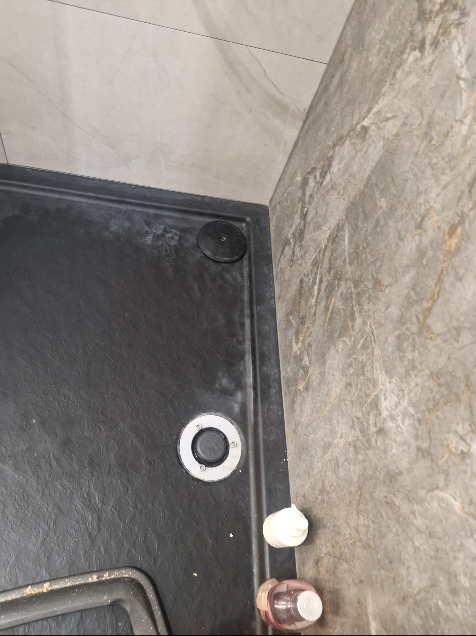 Czarny brodzik z odpływem i kosmetykami przy marmurowej ścianie. Widoczne ślady użytkowania, wymagające czyszczenia. Ujęcie z góry, bliski kadr.