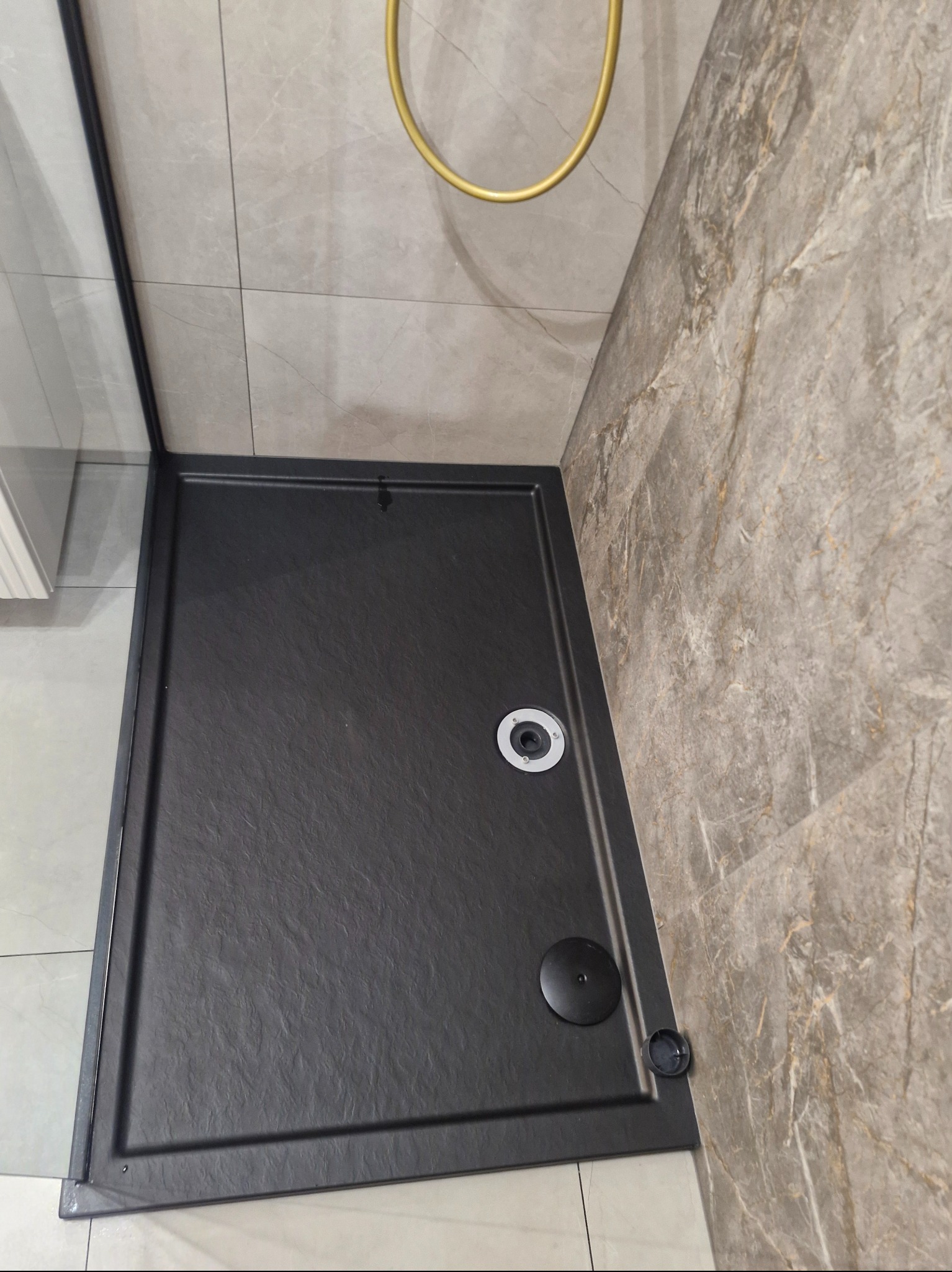 Nowoczesny, czarny brodzik kabiny prysznicowej z odpływem, otoczony marmurowymi płytkami. Złoty uchwyt prysznica w tle. Minimalistyczny design łazienki.