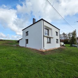 KG Elewacje - Biały, dwukondygnacyjny dom z ciemnymi oknami i dachem na zielonym trawniku pod błękitnym niebem z chmurami. Widoczne zabezpieczenia przy elewacji.