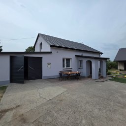 KG Elewacje - Szary dom jednorodzinny z ciemną dachówką i garażem z otwartymi drzwiami. Na podwórku stół z krzesłami. Widok z zewnątrz, po remoncie elewacji.