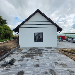 KG Elewacje - Biały dom z czarnym dachem i oknami, widok z przodu na nowo ułożony chodnik z kostki brukowej. W tle zieleń i pochmurne niebo.