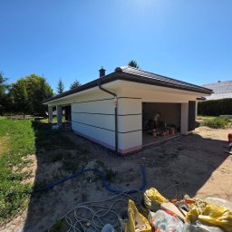 KG Elewacje - Nowoczesny, parterowy budynek z płaskim dachem, świeżo pomalowaną elewacją w odcieniach bieli i szarości, z widocznymi pracownikami w trakcie wykańczania detali.