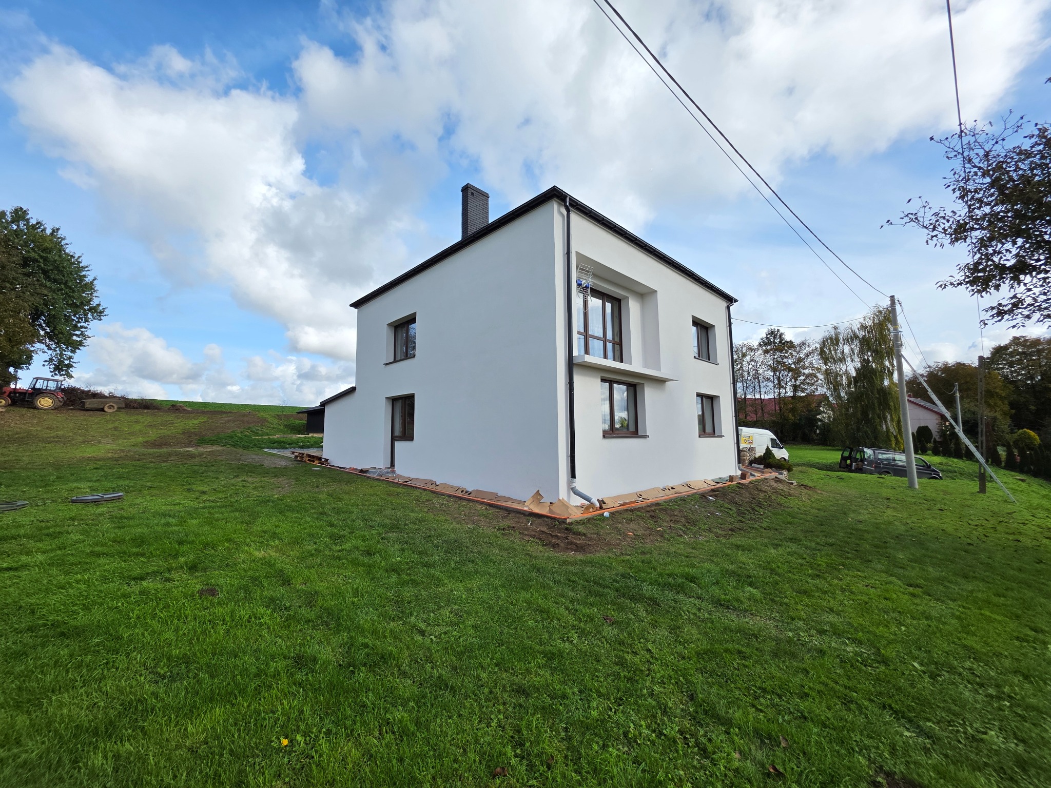 Biały, dwukondygnacyjny dom z ciemnymi oknami i dachem na zielonym trawniku pod błękitnym niebem z chmurami. Widoczne zabezpieczenia przy elewacji.