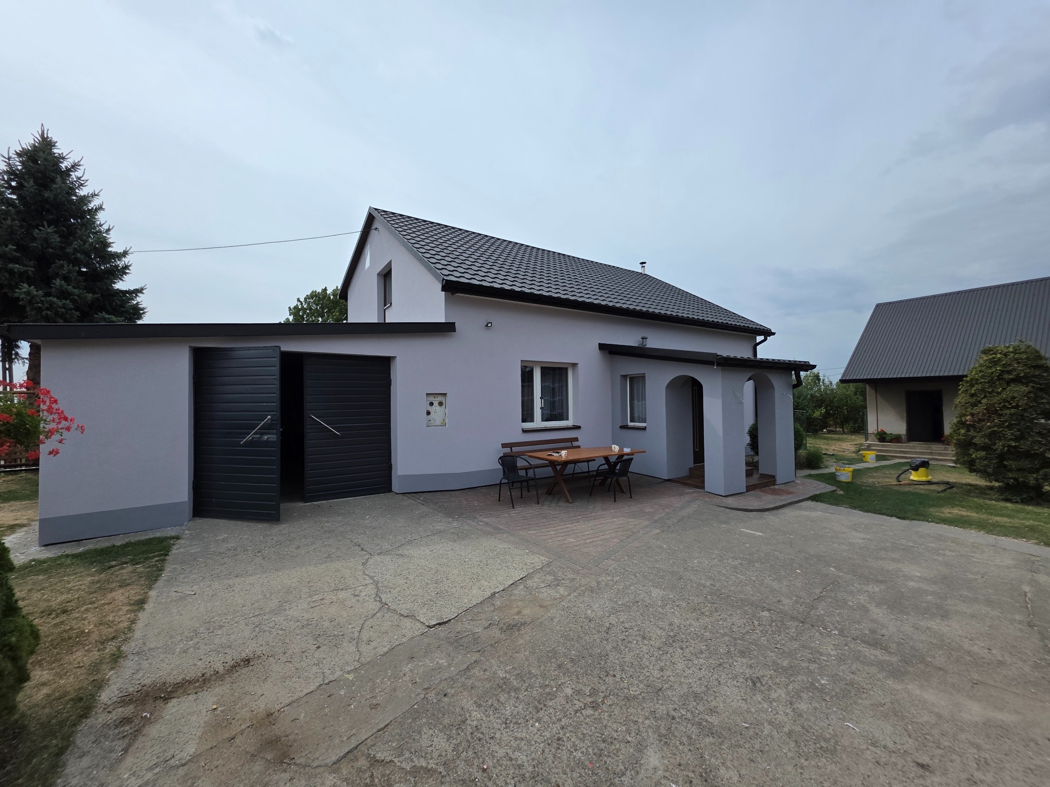 Szary dom jednorodzinny z ciemną dachówką i garażem z otwartymi drzwiami. Na podwórku stół z krzesłami. Widok z zewnątrz, po remoncie elewacji.