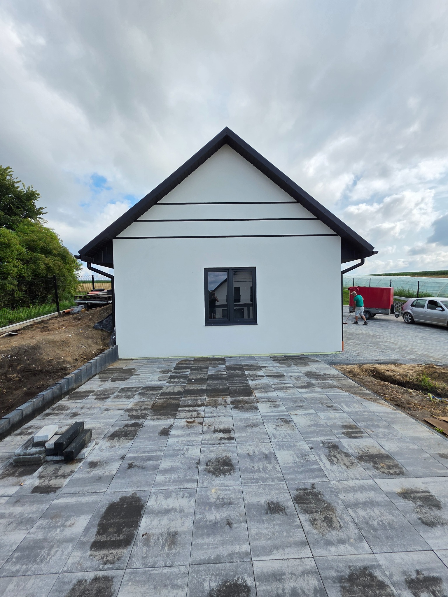 Biały dom z czarnym dachem i oknami, widok z przodu na nowo ułożony chodnik z kostki brukowej. W tle zieleń i pochmurne niebo.