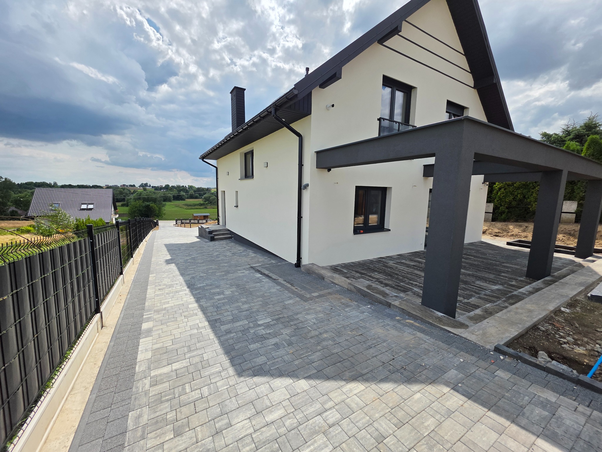 Nowoczesny dom jednorodzinny z jasną elewacją i ciemnymi elementami wykończeniowymi, widok z brukowanego podjazdu w słoneczny dzień.