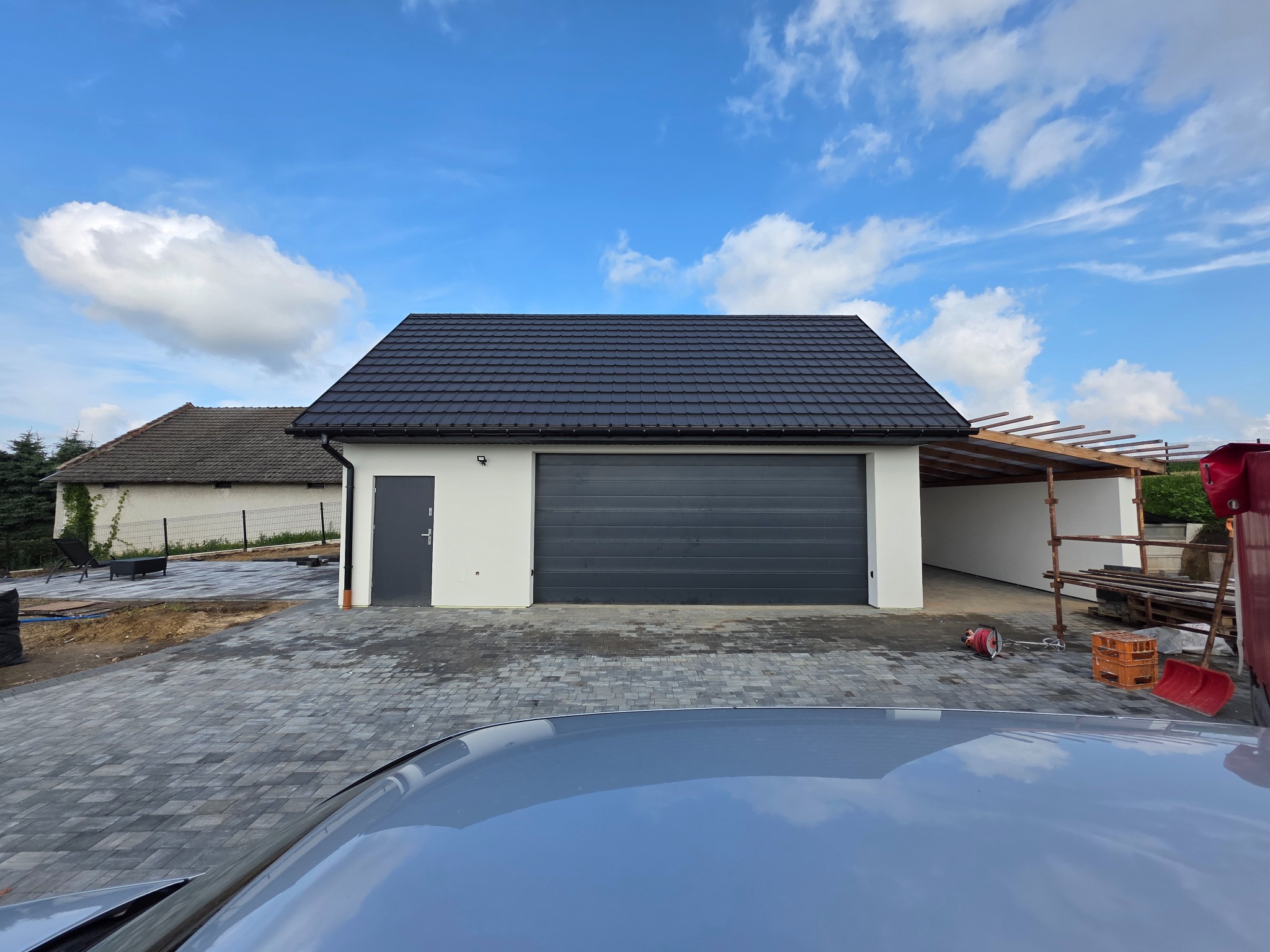 Nowoczesny garaż z szarą bramą segmentową i ciemną dachówką, świeżo wykończony, z kostką brukową na podjeździe. Widoczne elementy konstrukcji w tle.