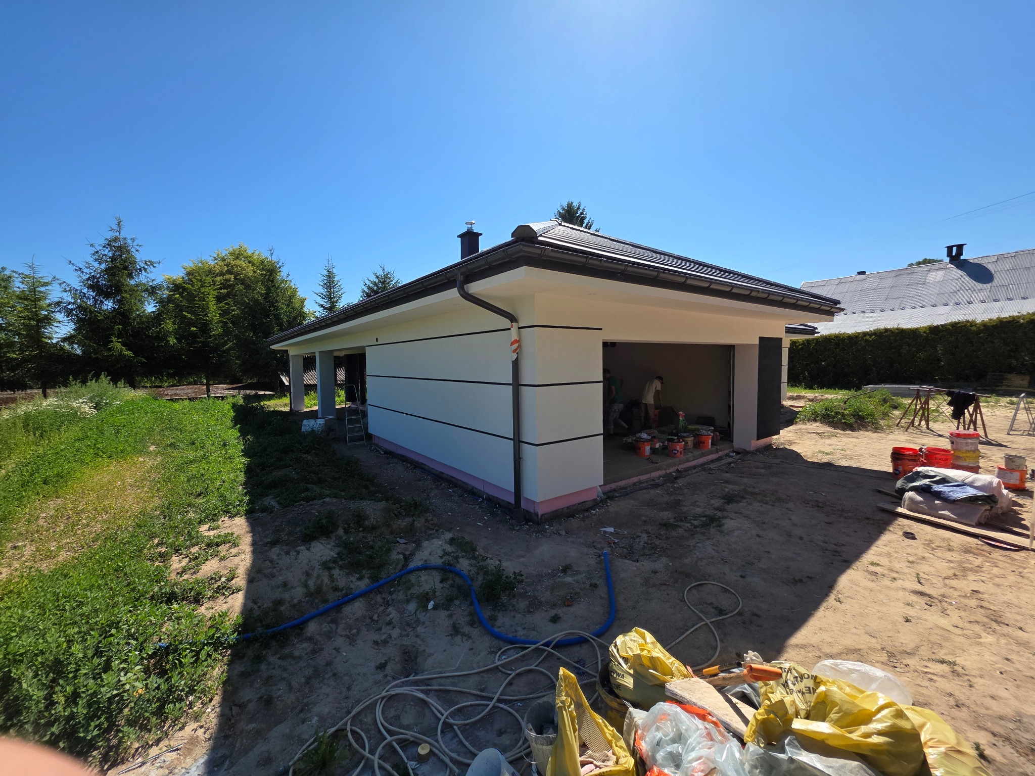 Nowoczesny, parterowy budynek z płaskim dachem, świeżo pomalowaną elewacją w odcieniach bieli i szarości, z widocznymi pracownikami w trakcie wykańczania detali.